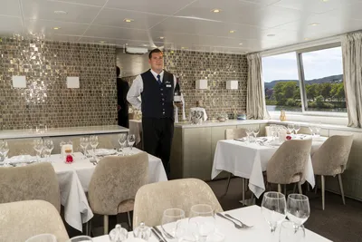 Flusskreuzfahrt Thurgau Maitre Der Maître in einem eleganten Restaurant auf einem Flusskreuzfahrtschiff von Thurgau Reisen. Der Raum ist geschmackvoll mit weißen Tischtüchern eingedeckt und mit Gläsern und Blumen dekoriert. Große Fenster bieten einen Blick auf die vorbeiziehende Landschaft, während der Maître bereit ist, die Gäste zu empfangen.