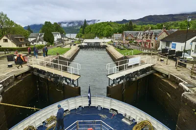 Flusskreuzfahrt Thurgau ReisenCaledonian Canal: fünf Schleusentreppen durch das Zentrum von F Blick auf eine Schleuse des Caledonian Canal, während ein Flusskreuzfahrtschiff von Thurgau Reisen einfahren möchte. Im Vordergrund ist das Schiff zu sehen, während die Schleusentore geöffnet sind. Im Hintergrund sind charmante Häuser und eine grüne Landschaft sichtbar, die die malerische Umgebung betonen.