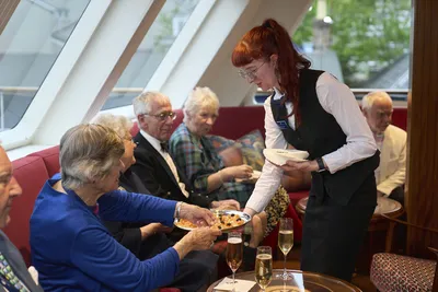 Flusskreuzfahrt Thurgau Apero Eine Kellnerin serviert Appetithäppchen während eines Aperitifs auf einem Flusskreuzfahrtschiff von Thurgau Reisen. Im Hintergrund sitzen mehrere Passagiere in gemütlicher Runde, genießen ihre Getränke und unterhalten sich. Die elegante Atmosphäre wird durch das stilvolle Interieur des Raumes und die ansprechende Präsentation der Speisen unterstrichen.