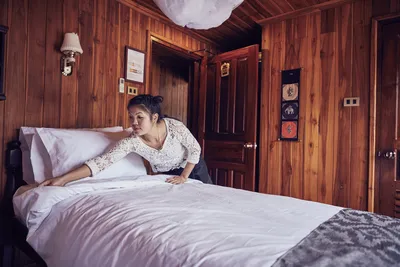 Flusskreuzfahrten Thurgau Mekong Pearl-ousekeeping Eine Mitarbeiterin des Housekeeping-Service auf dem Flusskreuzfahrtschiff Mekong Pearl von Thurgau Reisen macht das Bett in einer gemütlichen Kabine. Der Holzinterieur verleiht dem Raum eine warme Atmosphäre, während die Angestellte sorgfältig die Bettwäsche glättet.
