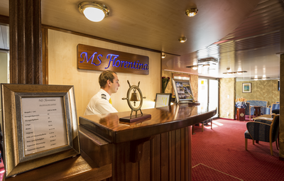 Hotelmanager Rezeption Flusskreuzfahrt MS Florentina-V1-P0 Der Empfangsbereich auf dem Flusskreuzfahrtschiff MS Florentina von Thurgau Reisen, mit dem Hotelmanager hinter dem Empfangstresen. Eine elegante Beschilderung mit dem Namen des Schiffes ist sichtbar, während im Hintergrund gemütliche Sitzgelegenheiten und eine freundliche Atmosphäre auf die Gäste warten.