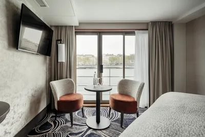 Thurgau Voyage Junior Suite Mitteldeck (ca.19 m²) mit franz. Balkon Innenansicht einer Kabine des Mitteldecks auf dem Flusskreuzfahrtschiff MS Voyage von Thurgau Reisen. Der Raum ist mit zwei gemütlichen Stühlen und einem kleinen Tisch eingerichtet, vor einem großen Fenster mit Blick auf den Fluss. Die moderne Einrichtung und die sanften Farben schaffen eine entspannte Atmosphäre.