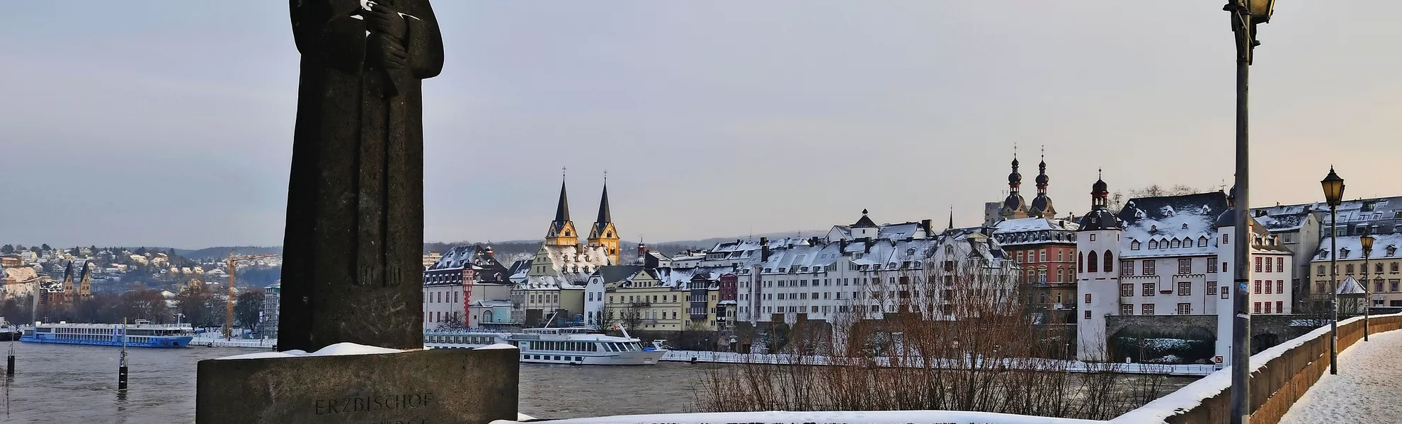 Blick auf Koblenz mit verschneiter Statue und Gebäuden entlang des Rheins im Winter.