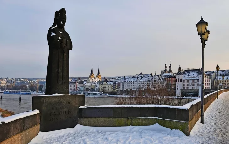 Blick auf Koblenz mit verschneiter Statue und Gebäuden entlang des Rheins im Winter.